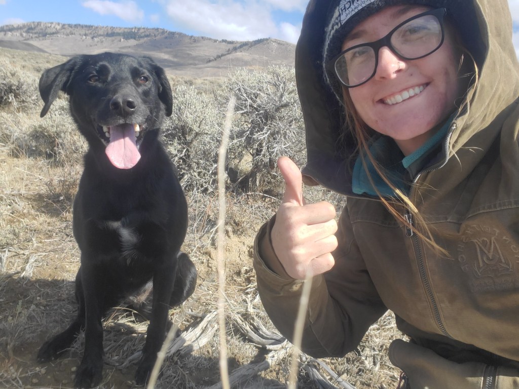 A black dog sits next to a smiling woman doing a thumbs up sign. They are in the middle of the sagebrush steppe.