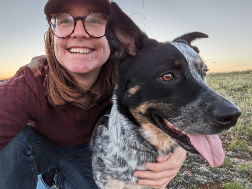 A woman smiles with her arm around a black and white dog. They are on an open prairie.